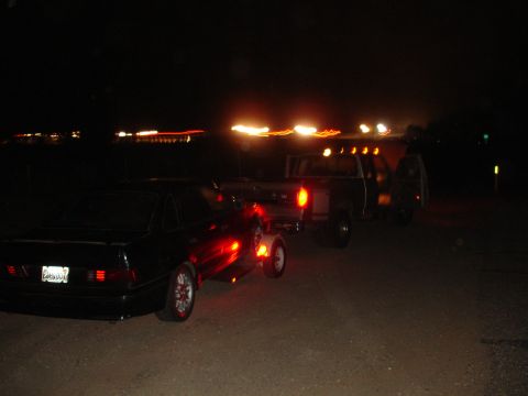 1989 Ford Taurus - A black sedan tows a trailer and a pickup truck in the dark.