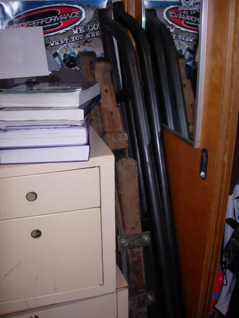 1985 Toyota Hilux - Leaf springs and exhaust pipes are stored indoors, leaning against a wall with posters and books nearby.