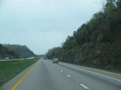1995 Ford C-350 (Centurion Conversions) - Cars travel on a multi-lane highway bordered by trees and rocky terrain under an overcast sky.