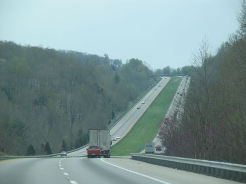 1995 Ford C-350 (Centurion Conversions) - Traffic moves along a highway surrounded by wooded hills on a cloudy day.