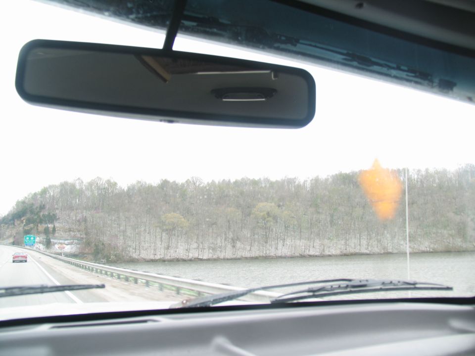 Interior view from a vehicle's windshield showing a highway, a body of water, and trees