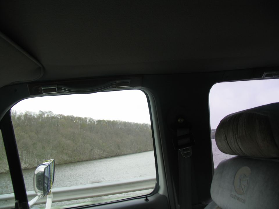 View from inside a vehicle, looking out the passenger side window at a body of water and trees. Seat headrest visible