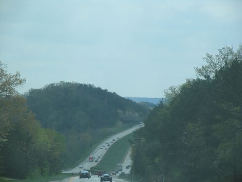 1995 Ford C-350 (Centurion Conversions) - Traffic moves along a highway surrounded by trees and hills under a cloudy sky.