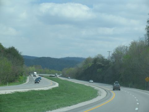 1995 Ford C-350 (Centurion Conversions) - Vehicles travel on a multi-lane highway surrounded by green hills and trees under a cloudy sky.