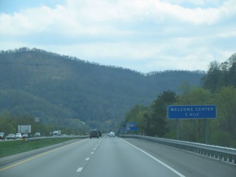 1995 Ford C-350 (Centurion Conversions) - View from a car on a highway approaching a welcome center, with forested mountains under a cloudy sky.