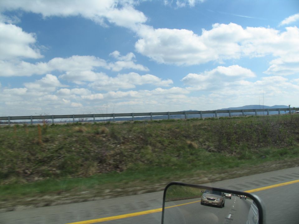 Car seen in side mirror on highway, with another car ahead. Guardrail and grassy embankment visible