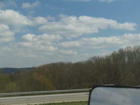 1995 Ford C-350 (Centurion Conversions) - A view from a vehicle's side mirror shows a highway and a tree-lined landscape under a cloudy sky.