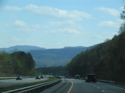 1995 Ford C-350 (Centurion Conversions) - A dark truck travels down a highway surrounded by rolling hills and mountains under a partly cloudy sky.