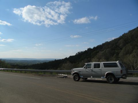 1995 Ford C-350 (Centurion Conversions) - A white extended cab pickup truck with a camper shell is parked on the side of a road overlooking a valley.