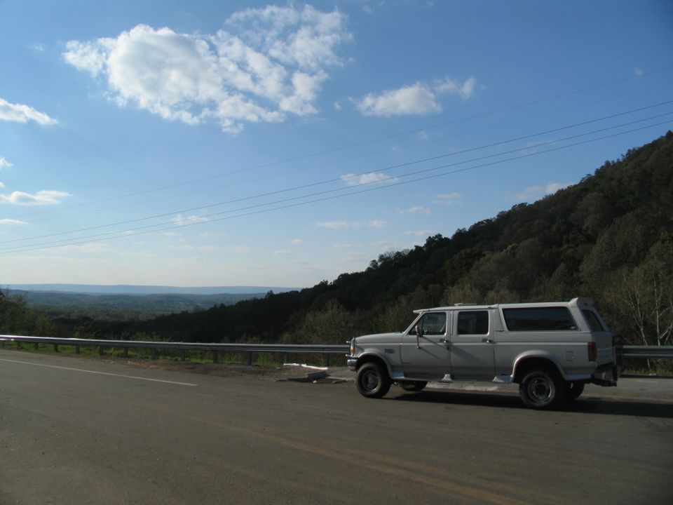 White extended cab pickup truck with a camper shell parked on the side of a road with a wooded hillside in the background