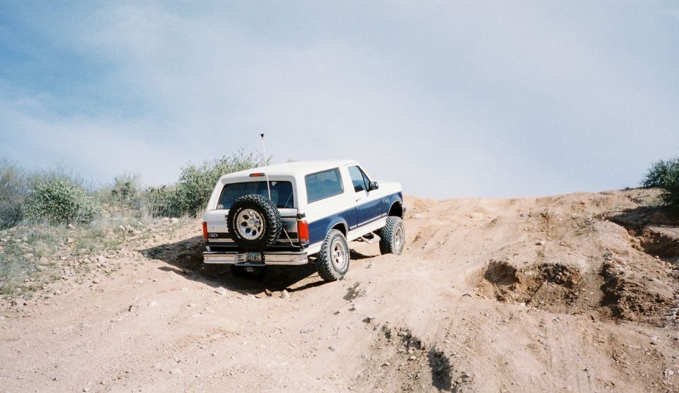 White and blue Ford Bronco climbing a dirt hill with a spare tire on the back