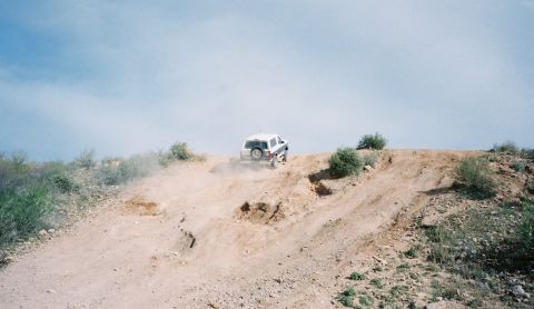 1996 Ford Bronco - A white SUV kicks up dust as it navigates a steep dirt incline under a clear blue sky.