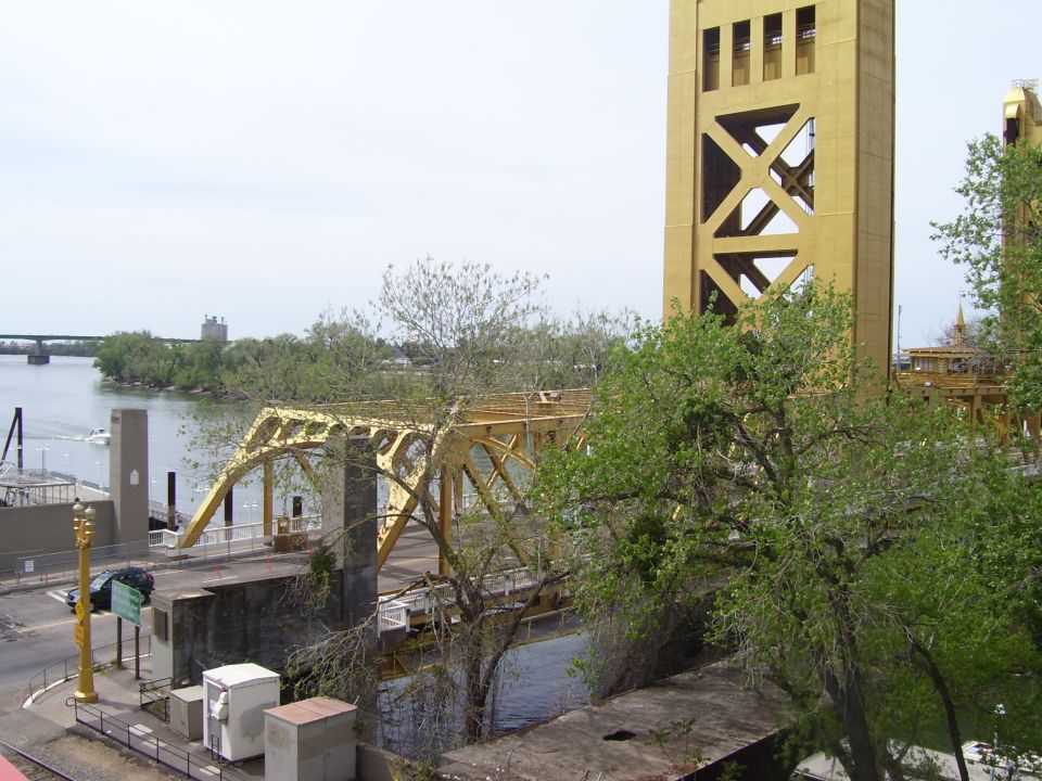 Yellow bridge tower and truss bridge over water, with trees and buildings in background