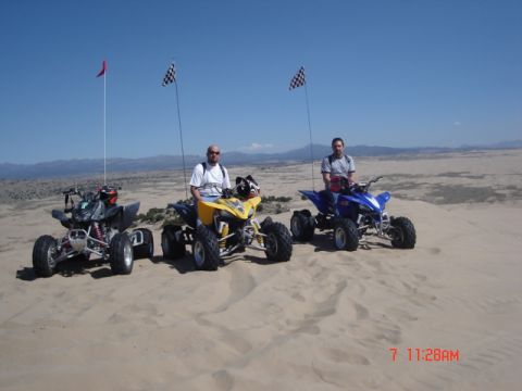 1981 Ford Bronco - Three ATVs are parked on sand dunes under a clear blue sky, with riders seated and flags attached to poles.