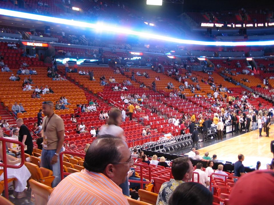 Spectators fill the red and orange seats of a basketball arena, with a court visible in the background