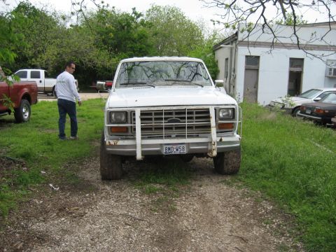 1986 Ford Bronco - A white Ford F-series truck is parked in a gravel lot with other vehicles and a building in the background.