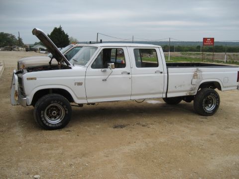 1986 Ford Bronco - White 1980-1986 Ford F-350 crew-cab long-bed pickup with hood raised, parked on a dirt lot; body shows rust and damage along the bed.