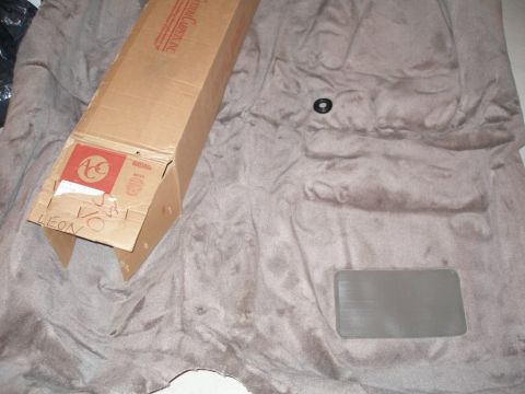2003 Ford Crown Victoria - A cardboard box sits on top of a piece of car carpet, with a small black circular object nearby.