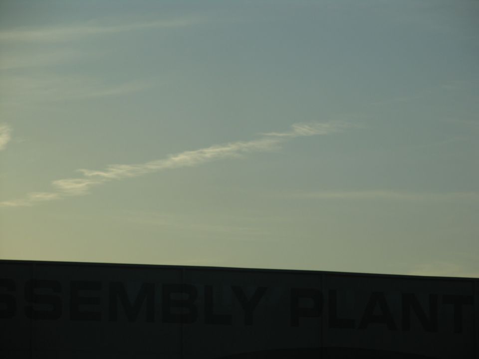 Building with the words ASSEMBLY PLANT visible on its side under a pale sky with a contrail