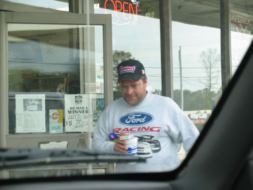 Man in Ford Racing sweatshirt and Summit Racing cap leaving a convenience store, seen through a windshield