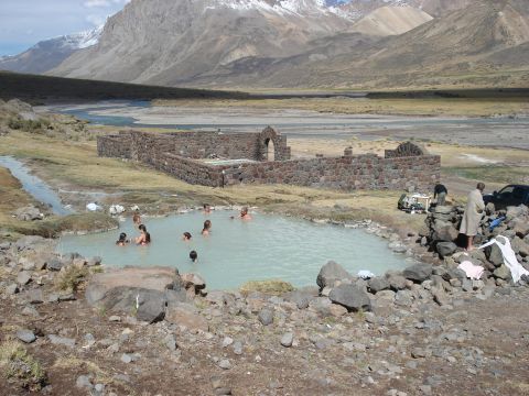1986 Ford Bronco - A remote hot spring with ancient stone structures and people enjoying the water amidst a vast, mountainous terrain.