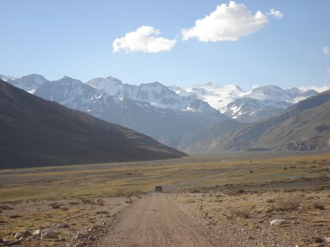 1986 Ford Bronco - A lone vehicle travels a dirt road through a vast valley towards snow-capped mountains under a blue sky.