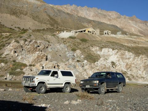 1986 Ford Bronco - Two SUVs parked in a remote, mountainous area with old industrial buildings on the hillside.