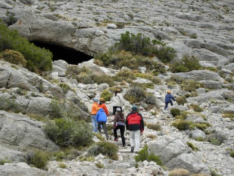 1986 Ford Bronco - A group of hikers ascends a rocky path towards a cave opening in a mountainous landscape.