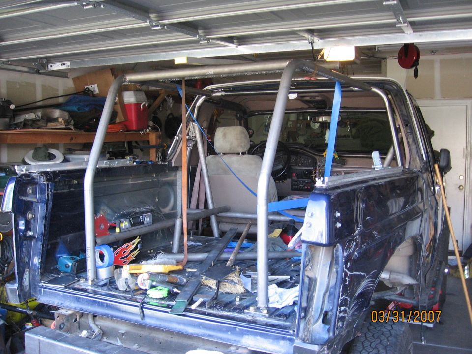 Blue SUV stripped to the floor pan in a garage with a mocked-up tube roll cage strapped in