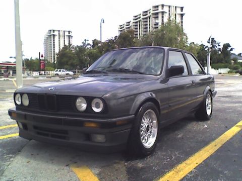 1989 BMW 3 Series - A dark gray 1989 BMW 3 Series coupe is parked in a lot with buildings in the background.