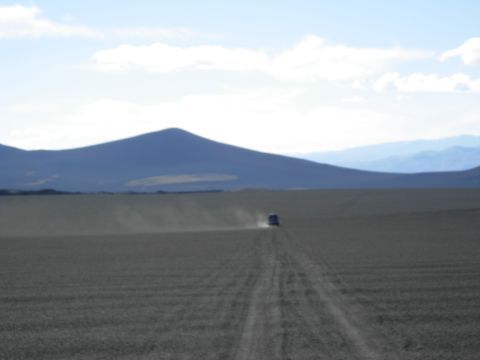 1986 Ford Bronco - A lone vehicle travels down a dusty track in a vast, open landscape under a blue sky.