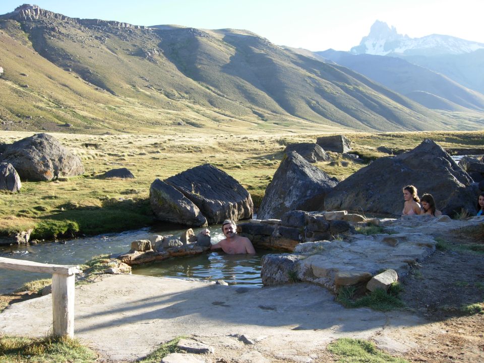 People relax in a rocky hot spring with mountains in the background. A watermark reads SUPERMOTORS.NET