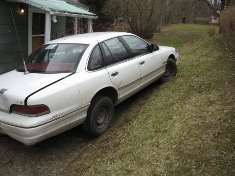 1992 Ford Crown Victoria - A white sedan sits parked on a grassy incline beside a house, showing signs of wear and tear.