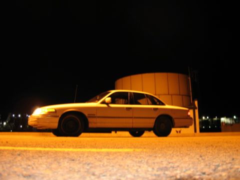 1992 Ford Crown Victoria - A yellow 1992 Ford Crown Victoria sedan is seen from the side at night, parked near a large cylindrical structure.