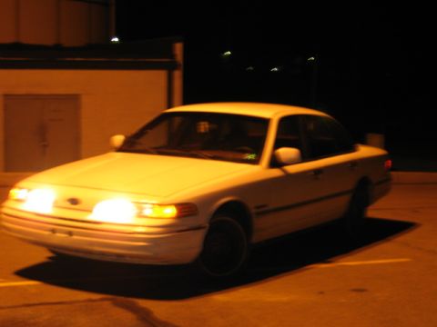 1992 Ford Crown Victoria - A white 1992 Ford Crown Victoria is captured from a 3q front angle at night, with its headlights illuminated.