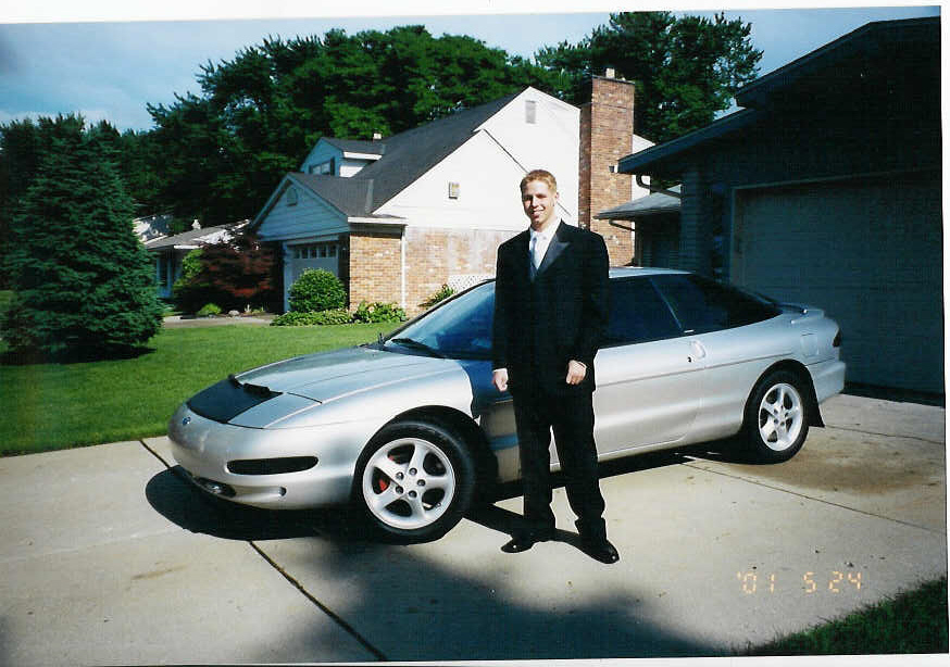 Silver Ford Probe with black hood bra in driveway, man in black suit standing beside it