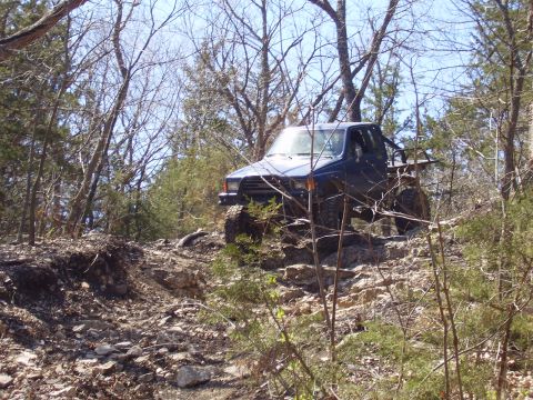 2007 Ford GT - A blue pickup truck is shown off-roading on a steep, rocky trail surrounded by trees.
