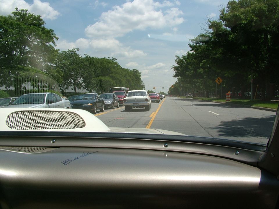 White 1959 sedan in traffic, rear view. Visible hood scoop and dashboard