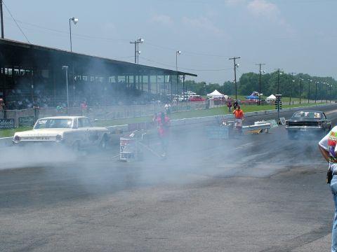 1972 Ford Ranchero - Two cars are racing on a drag strip, with one performing a burnout and kicking up smoke.