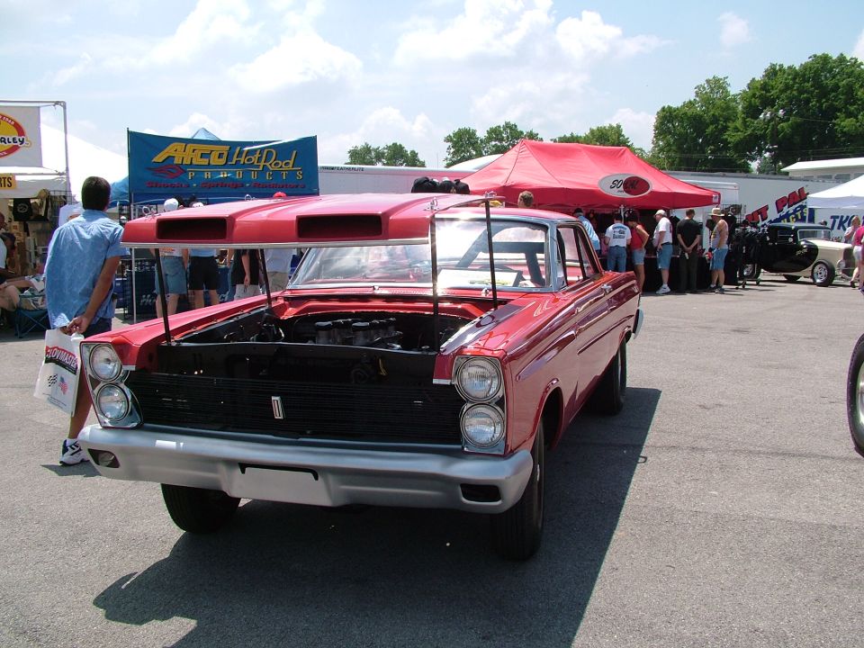 Red custom coupe with hood scoops and engine exposed. People milling around tents in background