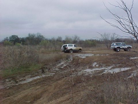 1978 Ford Bronco - Two SUVs navigate a challenging muddy terrain on an overcast day.