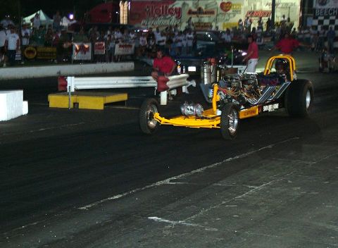 1972 Ford Ranchero - A dragster sits on the starting line of a race track at night, with spectators watching.