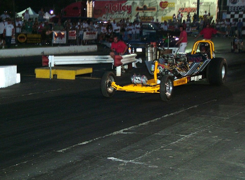 Yellow dragster with exposed engine on a race track, driver in seat, crowd in background