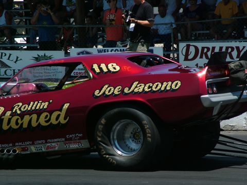 1972 Ford Ranchero - A red drag racing car with sponsor decals and driver's name is on the track with spectators in the background.