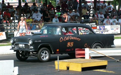 1972 Ford Ranchero - A black 1955 Chevrolet gasser drag car is on the track with spectators in the background.