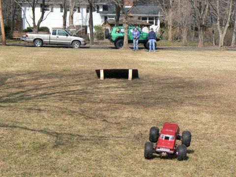 1994 Ford Bronco - A red remote control monster truck sits on a dry grassy field, with a jump ramp nearby and two full-size trucks parked in the background.