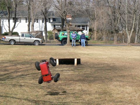1994 Ford Bronco - A red remote control car is captured mid-air over a grassy field, with two full-size vehicles and houses in the background.