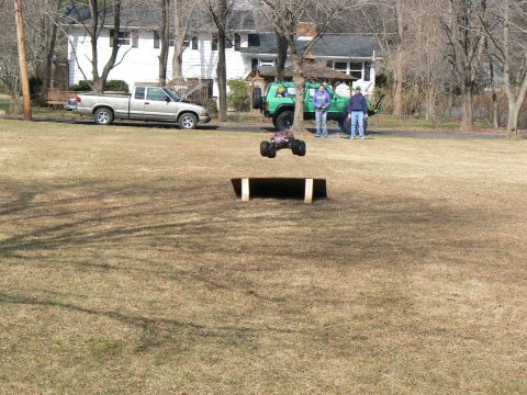 1994 Ford Bronco - A remote control monster truck jumps over a ramp in a grassy yard with other vehicles and people present.