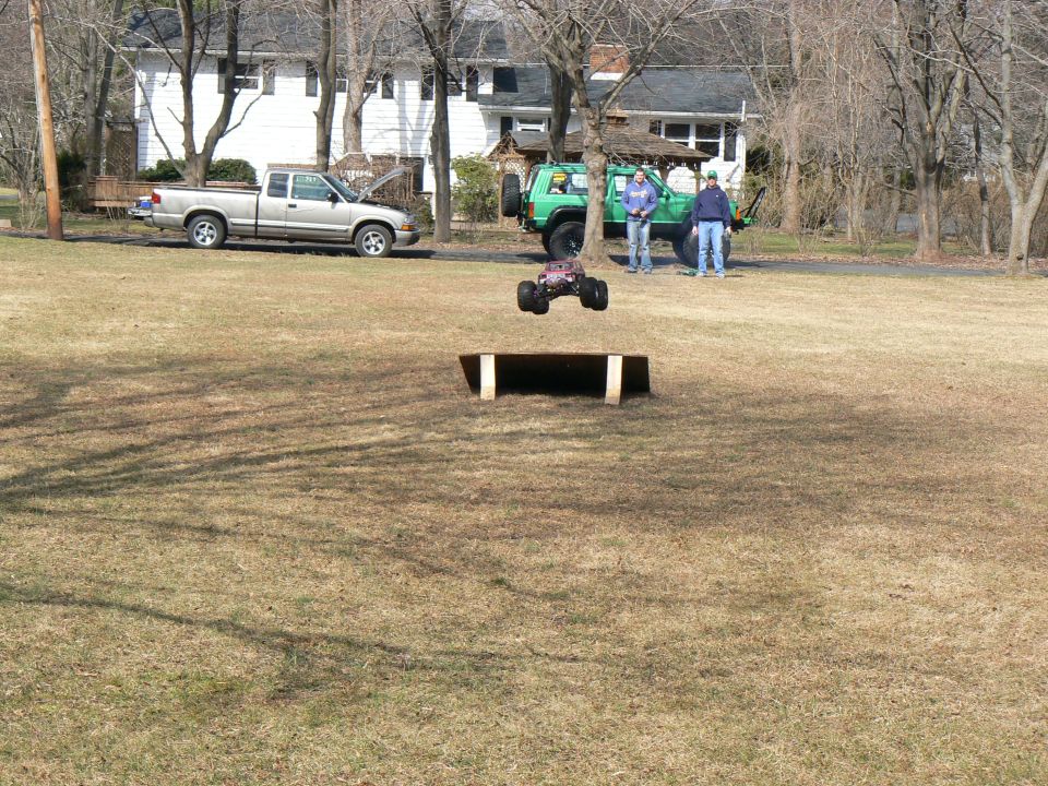 Remote control monster truck mid-air over a ramp with two people and two full-size vehicles in the background