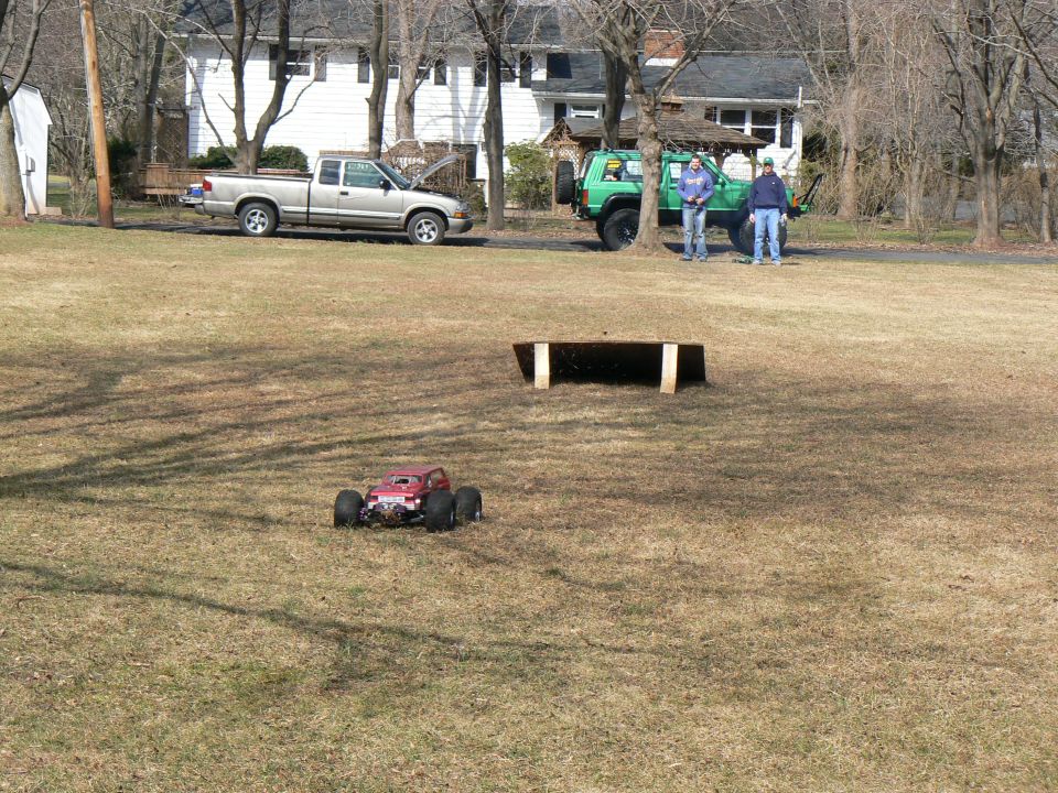 Red remote control monster truck on grass with a silver pickup truck and green Jeep in background